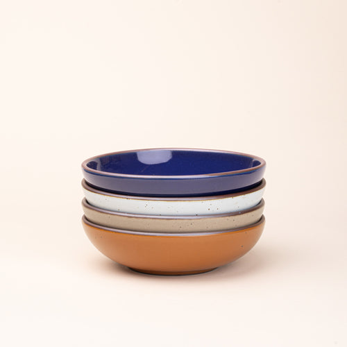 A stack of 4 dinner-sized shallow ceramic bowls in an earthy orange, glossy dark blue, white, and pale warm brown colors featuring iron speckles and an unglazed rim