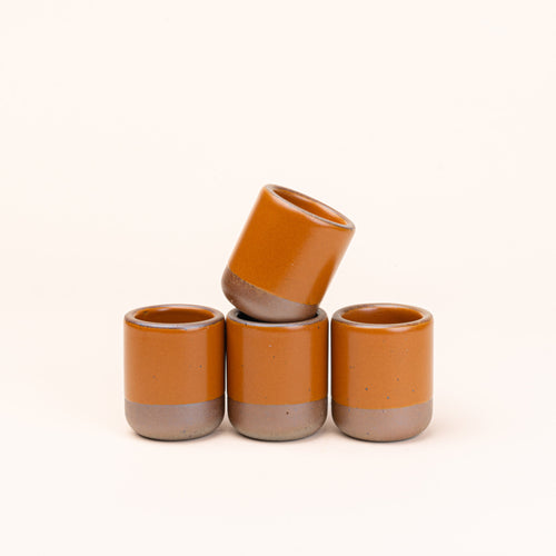A set of 4 small, short ceramic mug cups in an earthy orange color featuring iron speckles and unglazed rim and bottom base, stacked in a row of 3, with the 4th on top.