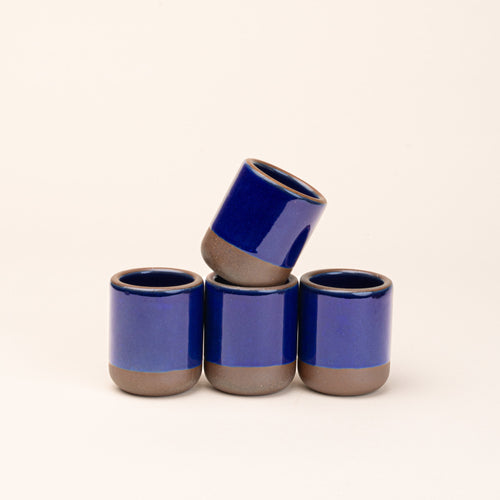 A set of 4 small, short ceramic mug cups in a glossy dark blue color featuring iron speckles and unglazed rim and bottom base, stacked in a row of 3, with the 4th on top.