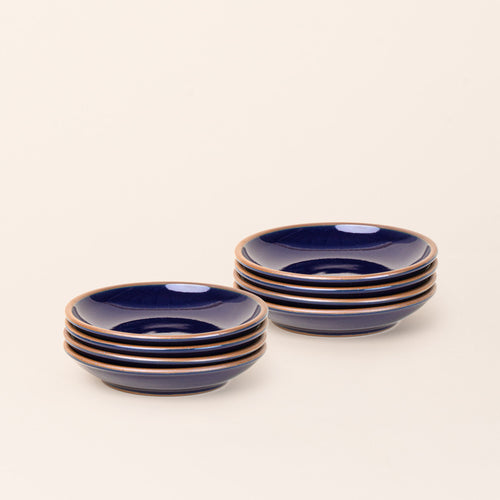 2 stacks of 4 small ceramic plates with a curved bowl edge in a glossy dark blue color featuring iron speckles and an unglazed rim.