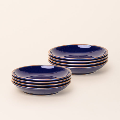 2 stacks of 4 medium ceramic plates with a curved bowl edge in a glossy dark blue color featuring iron speckles and an unglazed rim.
