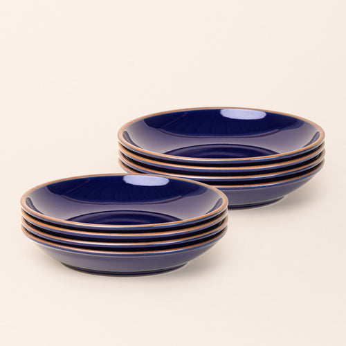 2 stacks of 4 large ceramic plates with a curved bowl edge in a glossy dark blue color featuring iron speckles and an unglazed rim.