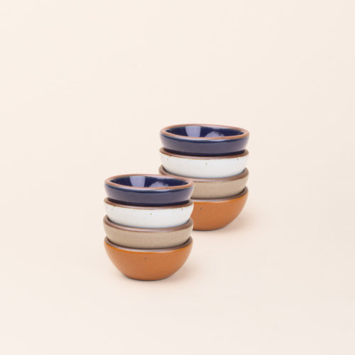 2 stacks of 4 tiny rounded ceramic bowls in an earthy orange, glossy dark blue, white, and warm pale brown colors featuring iron speckles and an unglazed rim