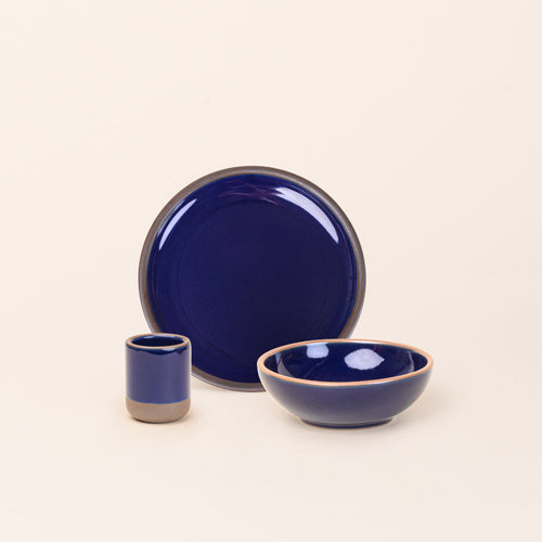 A small tiny cup, a medium sized plate, and a small breakfast bowl paired together in a glossy dark blue color featuring iron speckles.