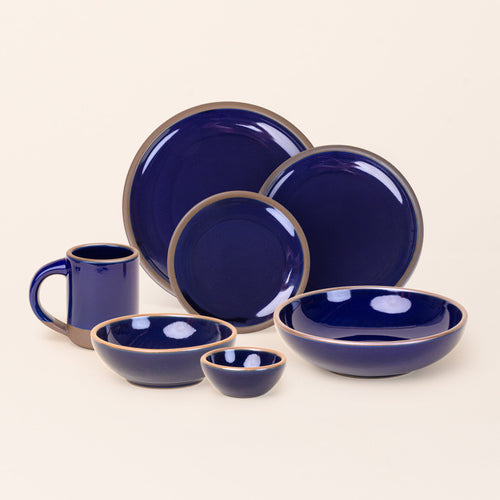 The Mug, bitty bowl, breakfast bowl, everyday bowl, cake plate, side plate and dinner plate paired together in a glossy dark blue color featuring iron speckles