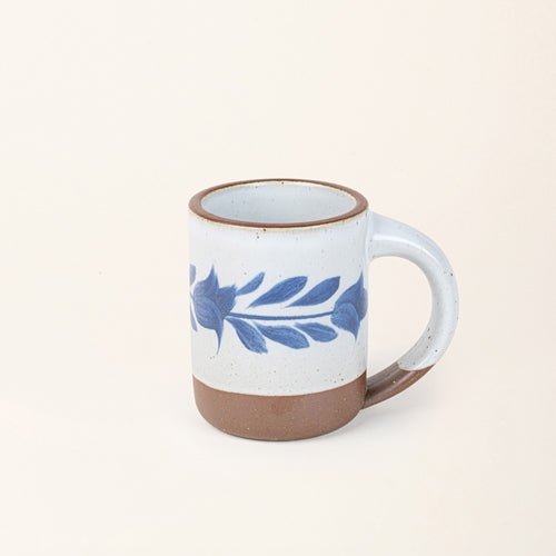 White ceramic mug with a hand-painted blue leaf motif and exposed clay base on a cream background.