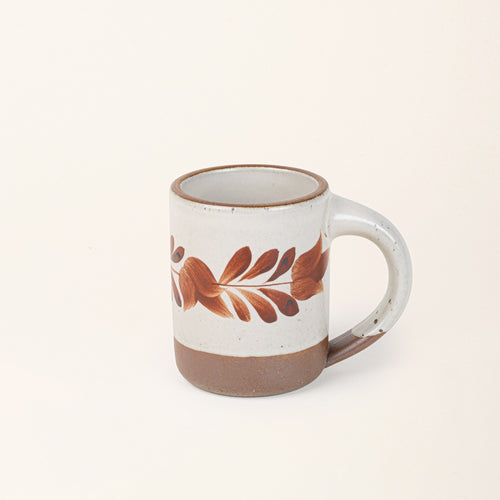 Warm off-white ceramic mug with a hand-painted terracotta leaf motif and exposed clay base on a cream background.