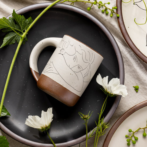 East Fork’s The Mug in a warm off white color with handle with a black decal of Henri Matisse’s Jeune femme la visage enfoui dans les bras, a bright blue, a line drawing of a young woman with face buried in her arms, on the body of the mug, styled on a black plate with flowers