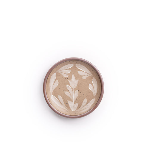 Small round ceramic plate with a hand-painted white flower and leaf design on a warm pale brown background with an exposed clay rim.