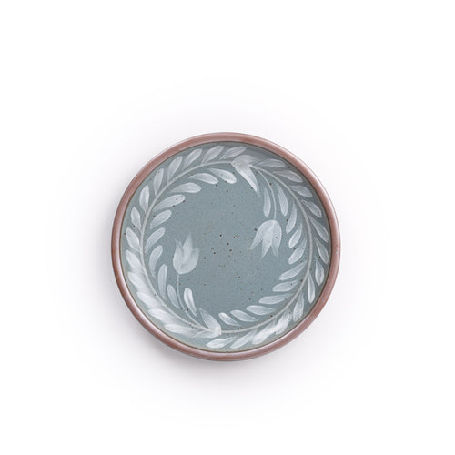 Side round ceramic plate with a hand-painted white leaf wreath and floral design on a blue-grey background with an exposed clay rim.