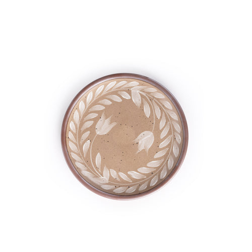 Side round ceramic plate with a hand-painted white leaf wreath and floral design on a warm pale brown background with an exposed clay rim.