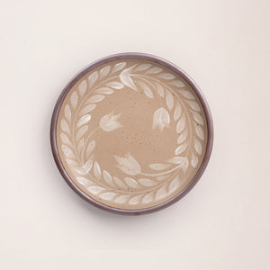 Dinner round ceramic plate with a hand-painted white leaf wreath and floral design on a warm pale brown background with an exposed clay rim.