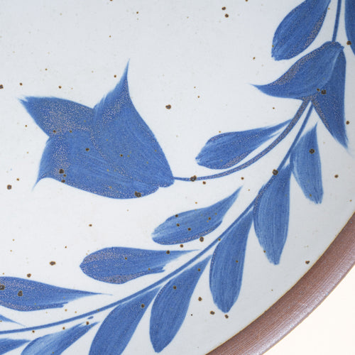 Closeup of a large round ceramic platter with a hand-painted blue leaf wreath and floral design on a white background with an exposed clay rim.