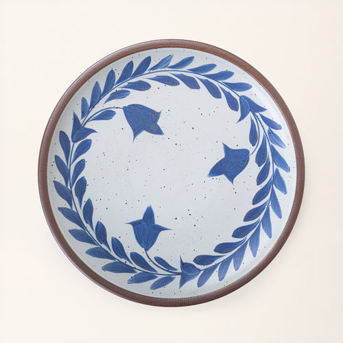 Large round ceramic platter with a hand-painted blue leaf wreath and floral design on a white background with an exposed clay rim.
