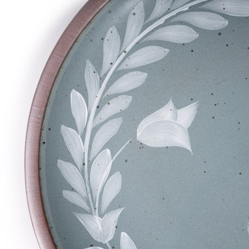 Closeup of a large round ceramic platter with a hand-painted white leaf wreath and floral design on a blue-grey background with an exposed clay rim.