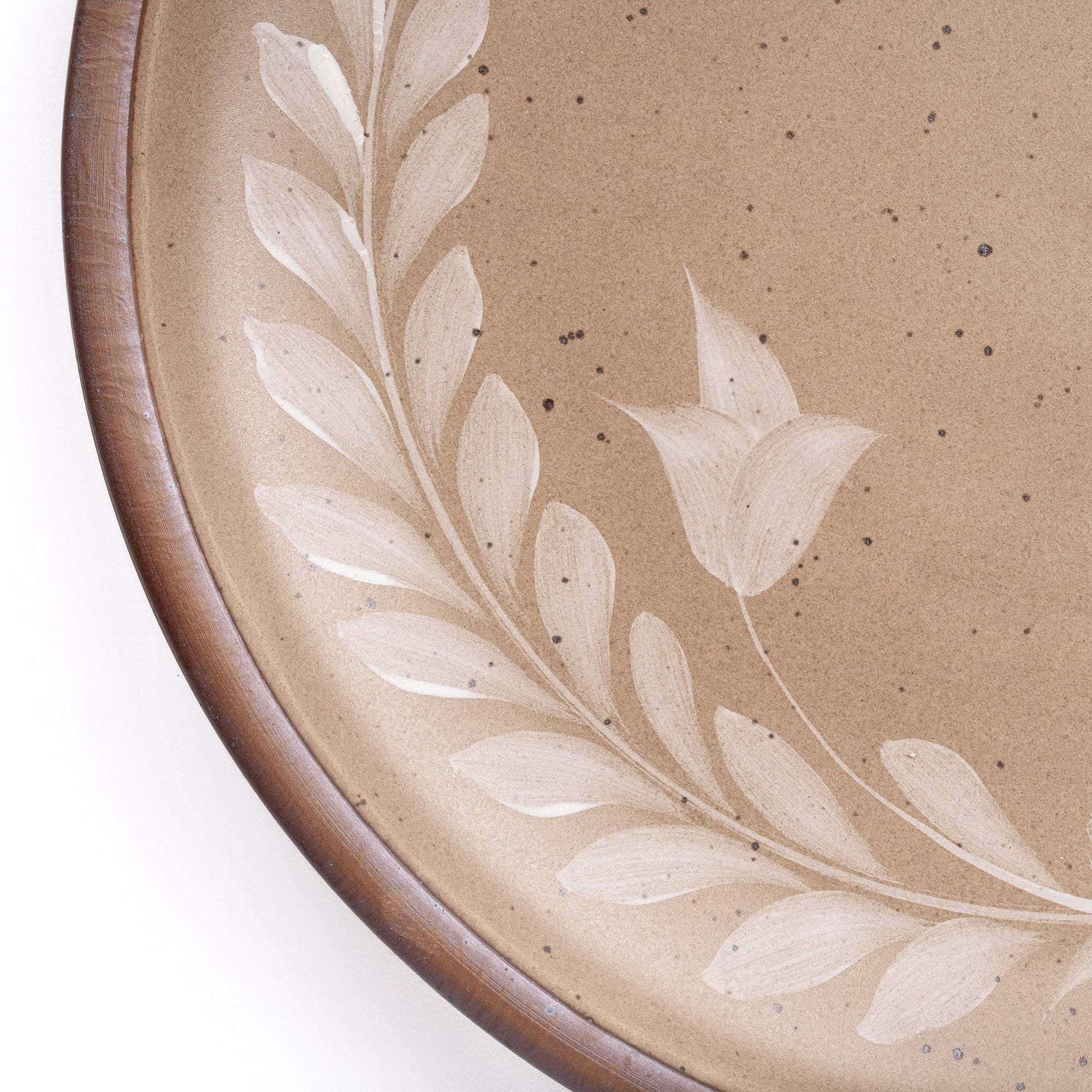 Closeup of a large round ceramic platter with a hand-painted white leaf wreath and floral design on a warm pale brown background with an exposed clay rim.