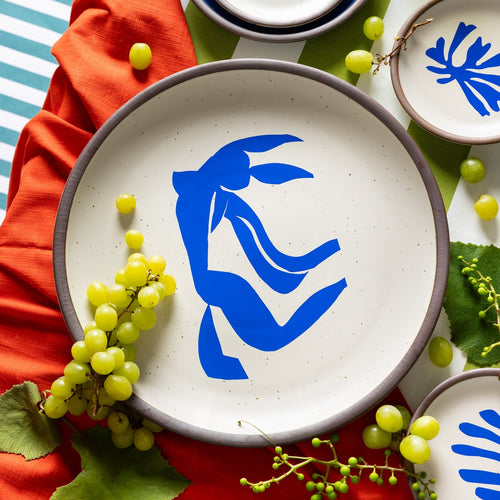 A styled tabletop photo of A large serving platter in a warm off-white color with unglazed rim with a blue decal of the artwork, La Chevelure, from Henri Matisse, with other Matisse cake plates, grapes, and colorful linens.