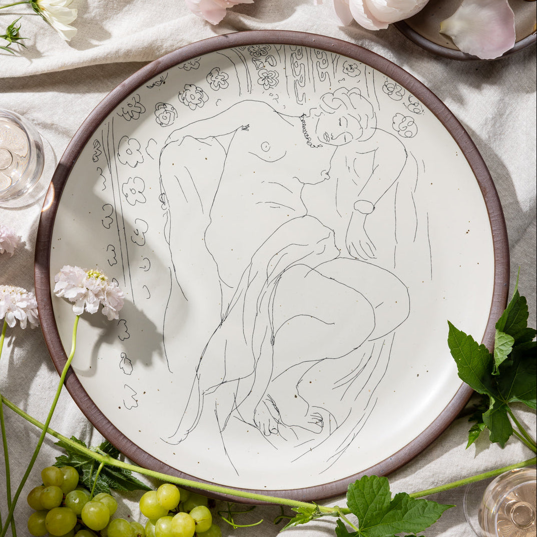 A large serving platter in a warm off-white color with unglazed rim with a black decal of the artwork, Figure endormie sur fond moucharabieh, from Henri Matisse, styled on a tablecloth with grapes and flowers.