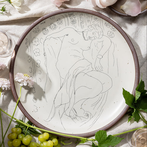 A large serving platter in a warm off-white color with unglazed rim with a black decal of the artwork, Figure endormie sur fond moucharabieh, from Henri Matisse, styled on a tablecloth with grapes and flowers.