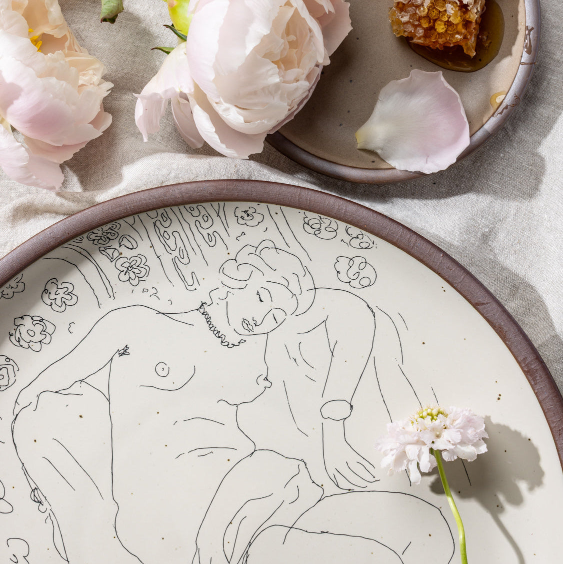 Closeup of a large serving platter in a warm off-white color with unglazed rim with a black decal of the artwork, Figure endormie sur fond moucharabieh, from Henri Matisse, styled on a tablecloth with grapes and flowers.