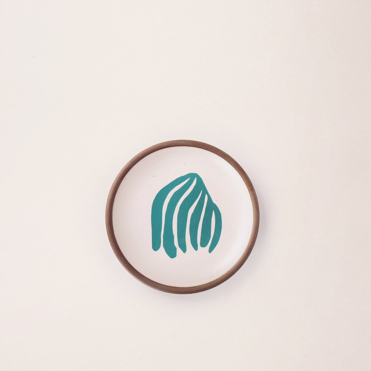 A ceramic dessert sized plate in a warm off-white color with unglazed rim, a teal frond-like cut-outs from Henri Matisse’s La Perruche et la sirène.