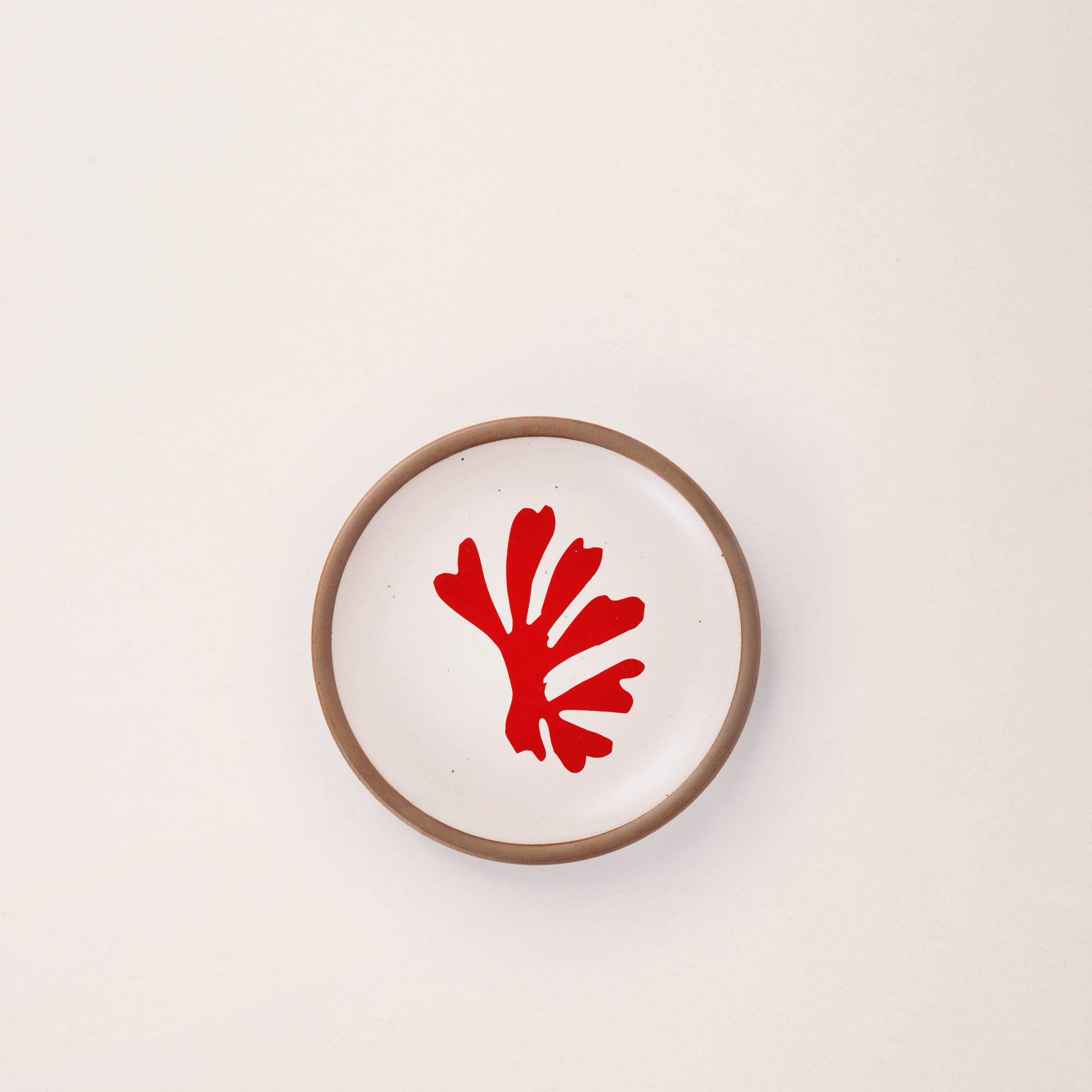 A ceramic dessert sized plate in a warm off-white color with unglazed rim, a red frond-like cut-outs from Henri Matisse’s La Perruche et la sirène.