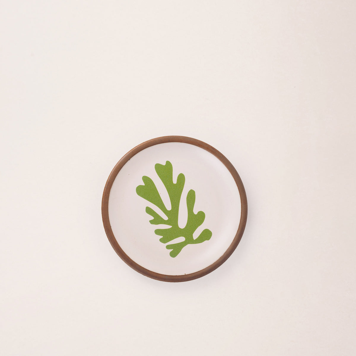 A ceramic dessert sized plate in a warm off-white color with unglazed rim, a green frond-like cut-outs from Henri Matisse’s La Perruche et la sirène.
