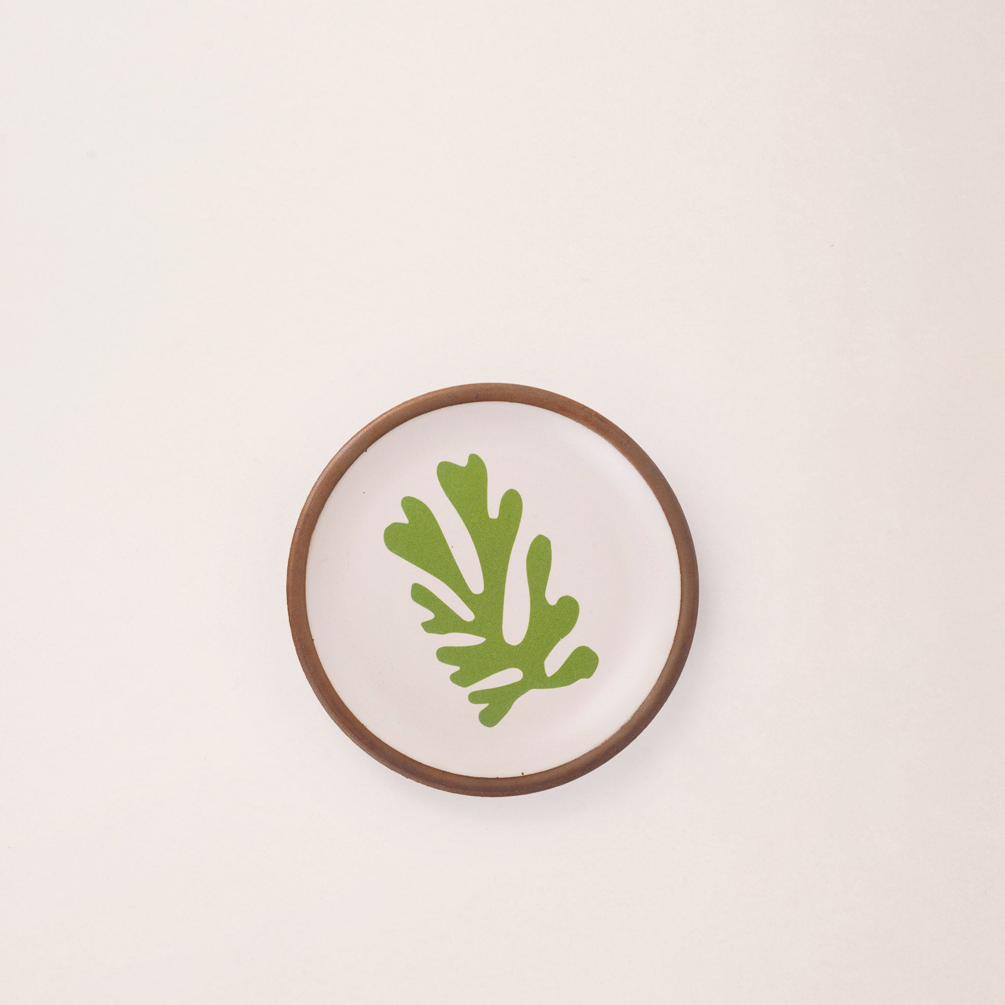 A ceramic dessert sized plate in a warm off-white color with unglazed rim, a green frond-like cut-outs from Henri Matisse’s La Perruche et la sirène.