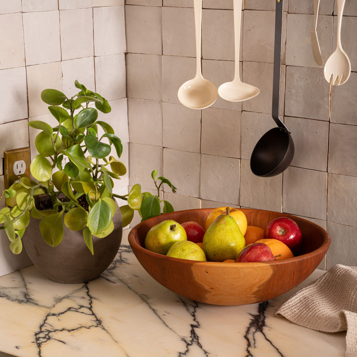 Cherry wood fruit bowl filled with pears, apples, and oranges on a marble countertop beside a leafy potted plant and hanging kitchen utensils.