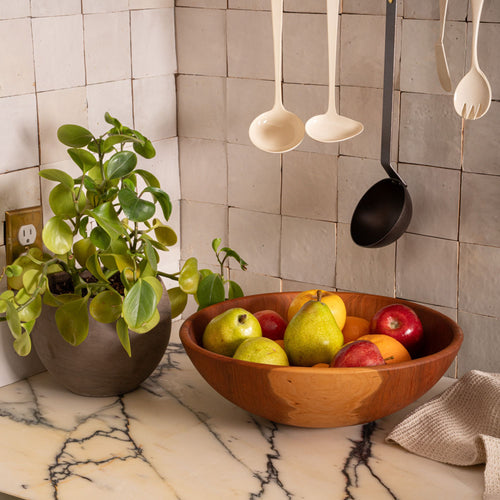 Cherry wood fruit bowl filled with pears, apples, and oranges on a marble countertop beside a leafy potted plant and hanging kitchen utensils.