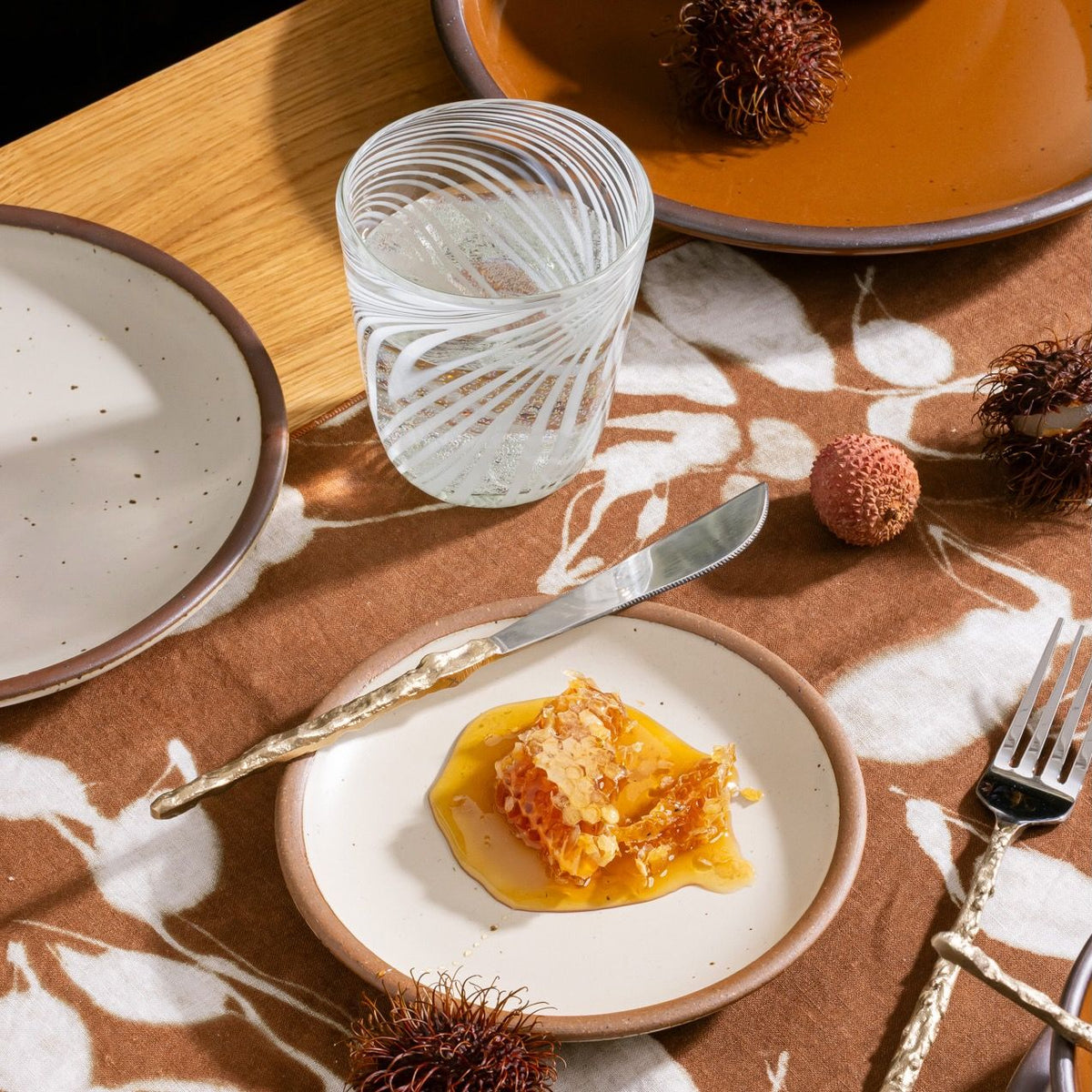 Honeycomb with syrup on a ceramic plate beside a patterned glass of water, rambutan fruits, and brown dinnerware on a floral cloth.