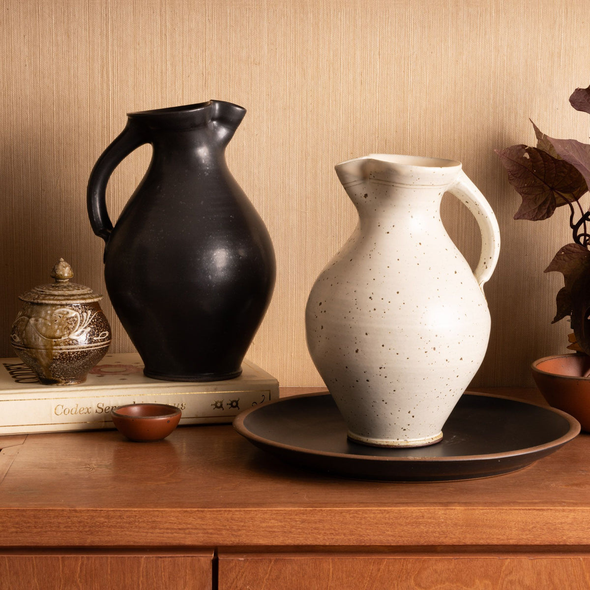 Black and cream ceramic pitchers on a wooden surface, styled with books, bowls, and foliage against a neutral wall.