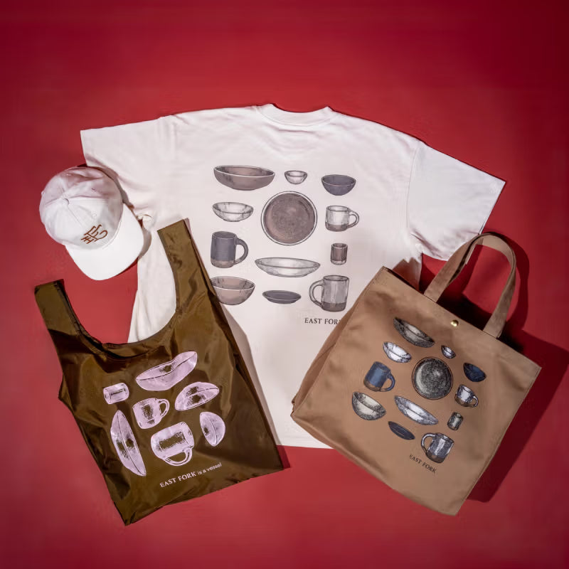 Overhead view of an East Fork tee with ceramic mugs, plates, and mug illustration, with a tote bag, a nylon bag, and a baseball hat - all against a red background.