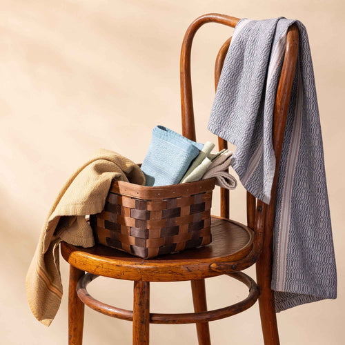Small rectangular woven wood basket in two-tone brown with an open top and reinforced rim, shown on a chair styled with a tea towels.