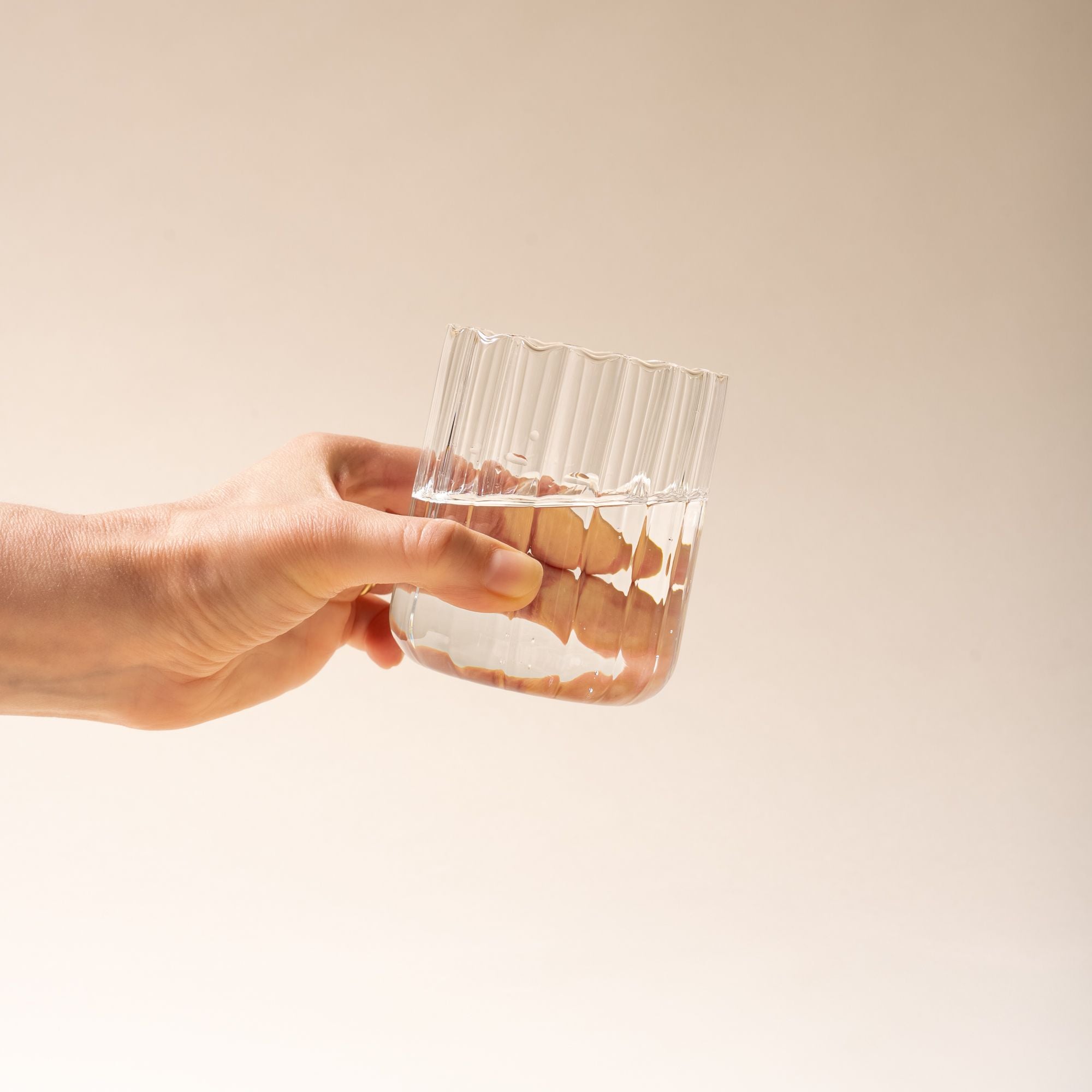 Hand holding a clear glass of water, half-filled, with vertical ridges on a neutral background.