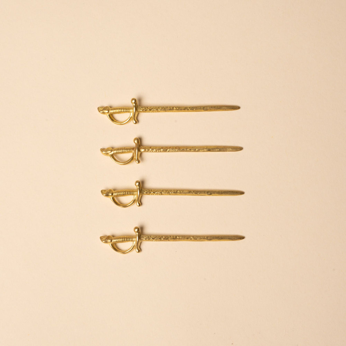 Set of four brass cocktail picks shaped like miniature swords, laid in a row on a neutral background.