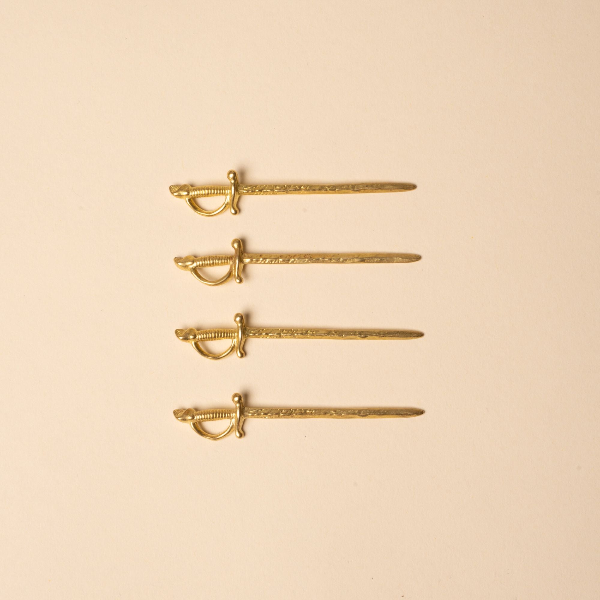 Set of four brass cocktail picks shaped like miniature swords, laid in a row on a neutral background.