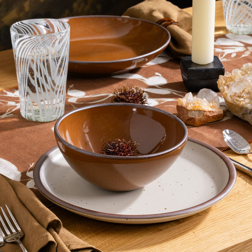 Glossy caramel brown ceramic bowl and matte placed on a matching off-white plate with rim on a table with silverware and drinking glass.