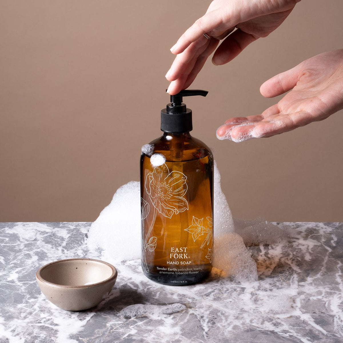 A brown glass bottle of East Fork Hand Soap labeled “Tender Earth” with hands dispensing with soap bubbles and a small tiny bowl.