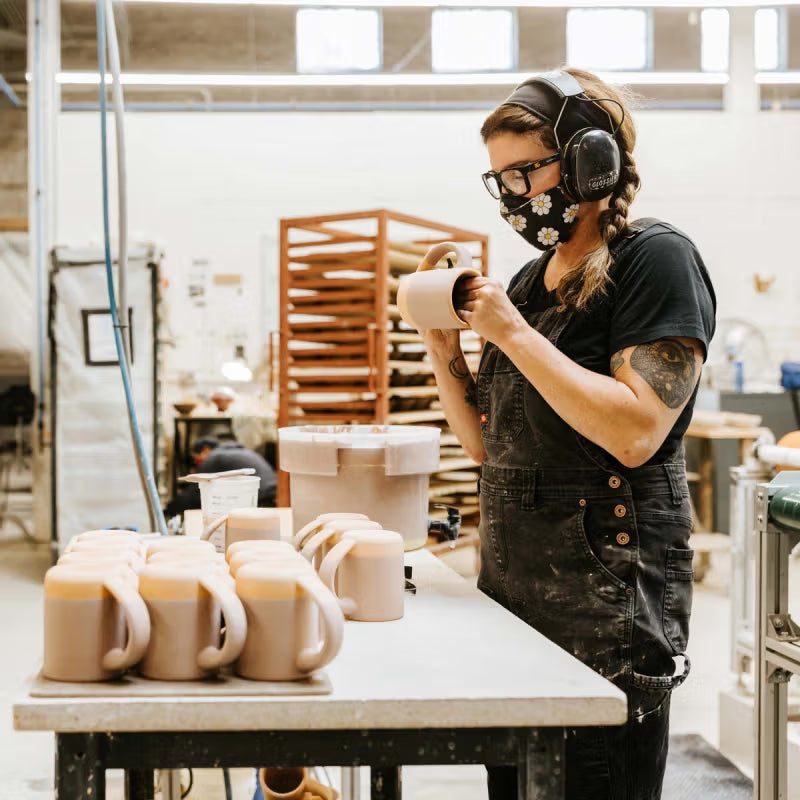 A person wearing safety headphones and a mask is working on mug and standing in front of a table of unglazed mugs.