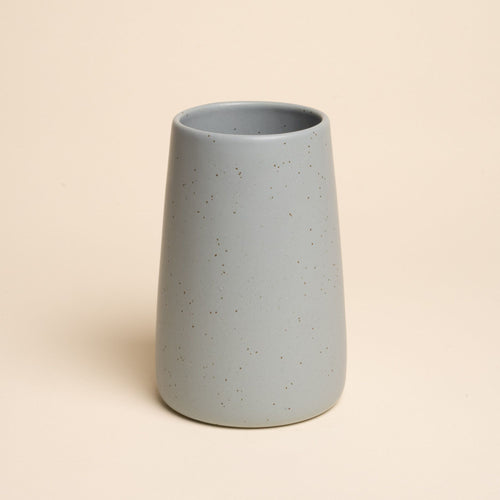 A large minimalist blue-grey tumbler vase with subtle iron speckles stands centered on a soft beige background
