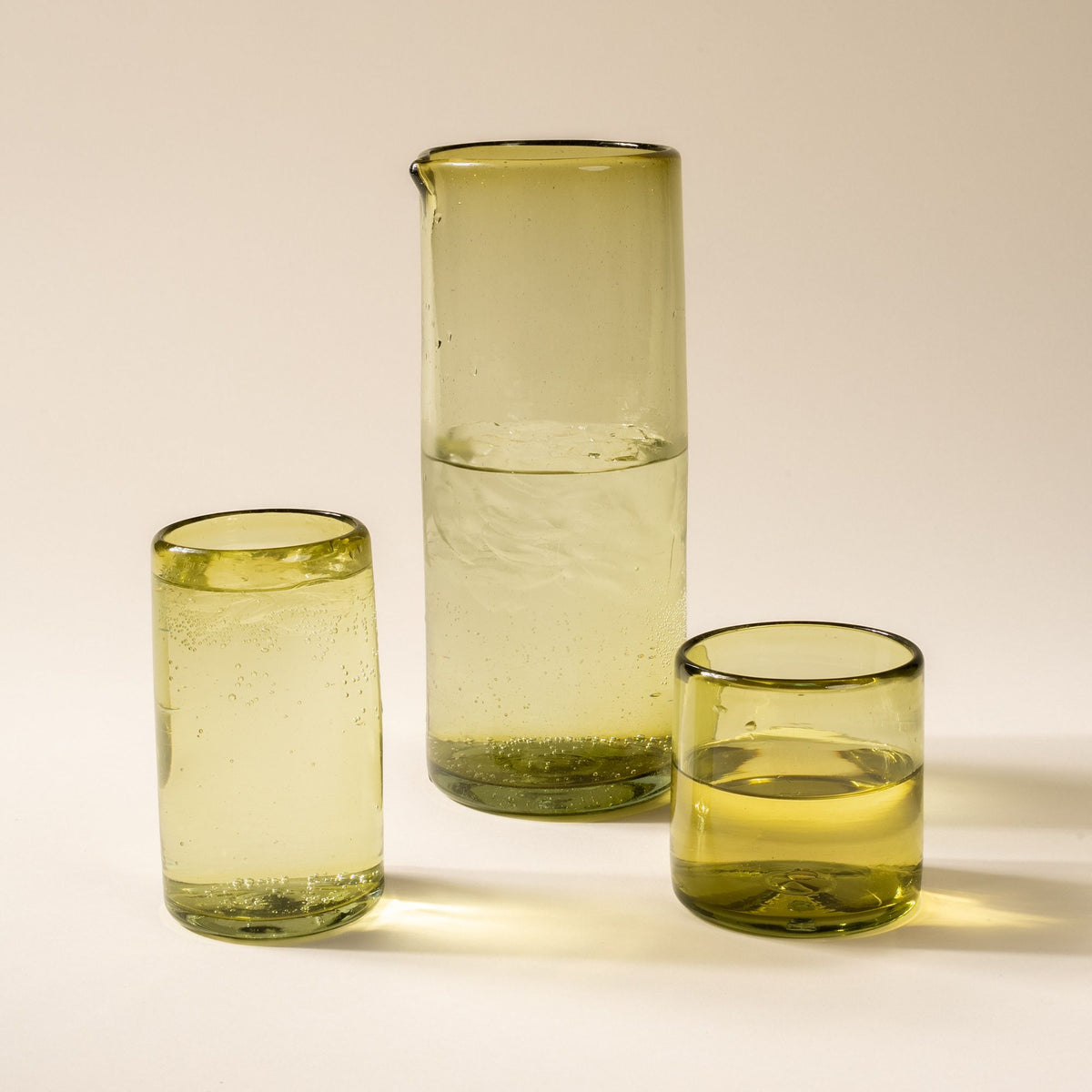 A tall tumbler, large pitcher, and short tumbler, of transparent glassware in an olive green color, all filled with drinks.