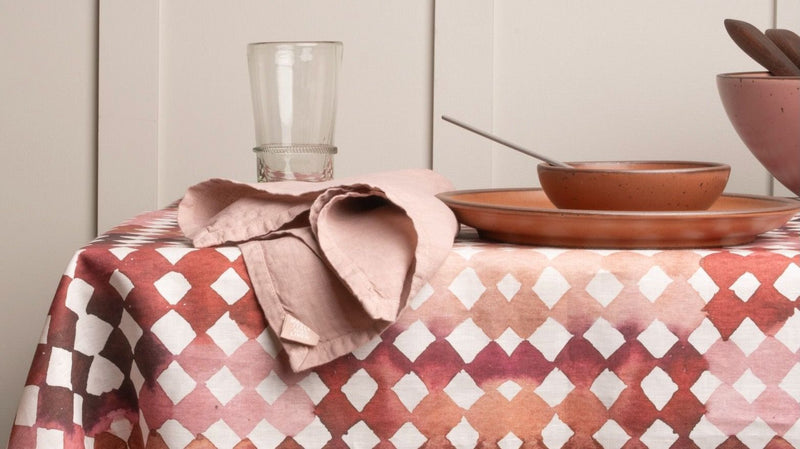 Table set with pink ceramic dishware on a watercolor diamond patterned tablecloth, with a wooden chair and linen napkin nearby.