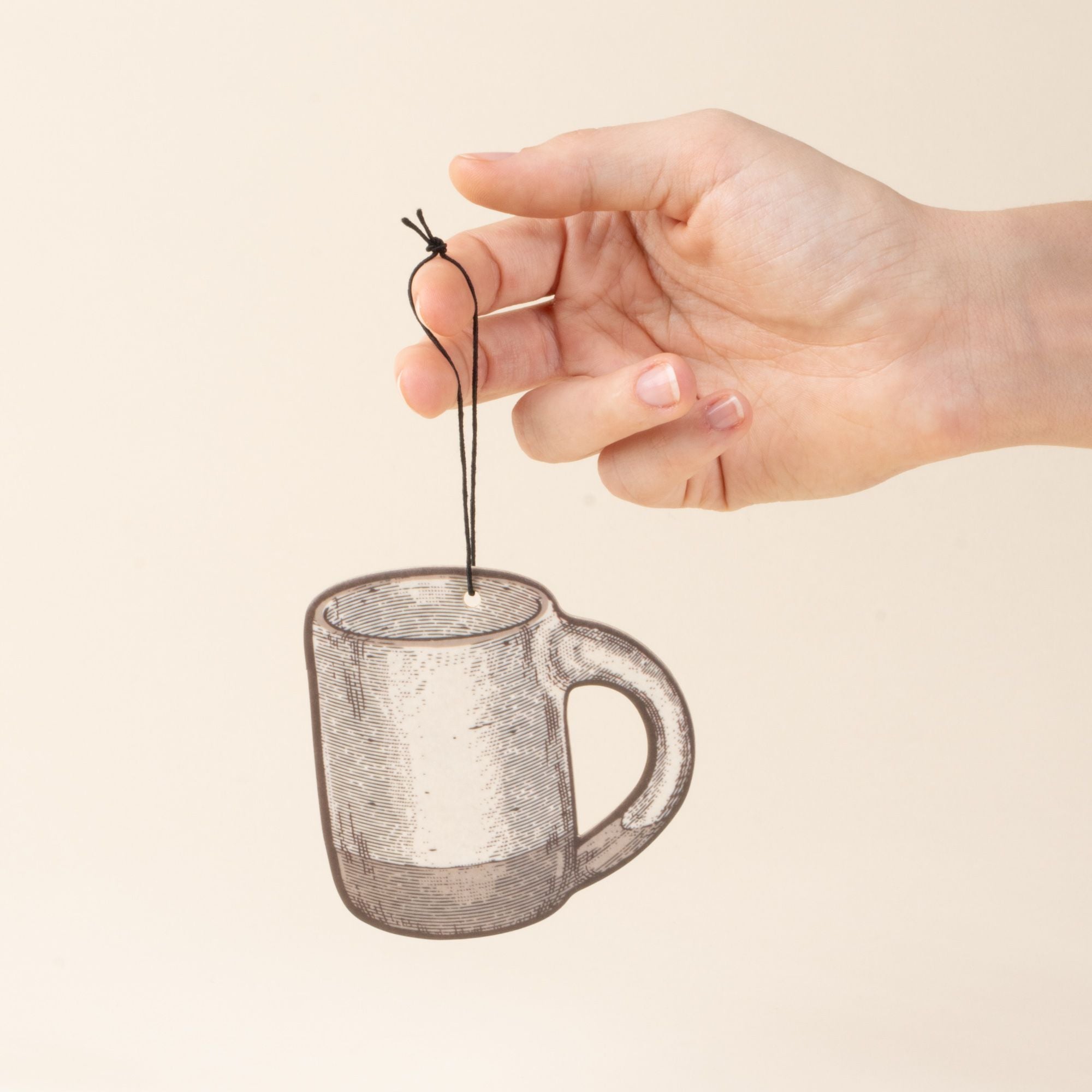 A hand holding an illustrated mug-shaped air freshener in a cool white color with a black hanging string against a light cream background.