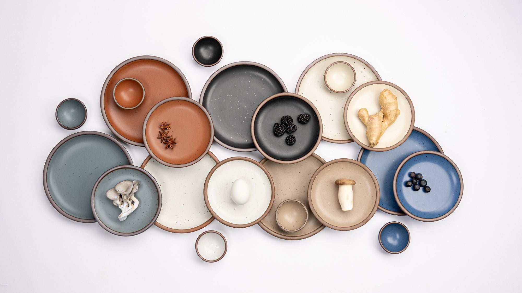 An overhead view of ceramic plates and bowls artfully arranged in East Fork core colors: Heron, Amaro, Eggshell, Black Mountain, Morel, Panna Cotta, and Blue Ridge
