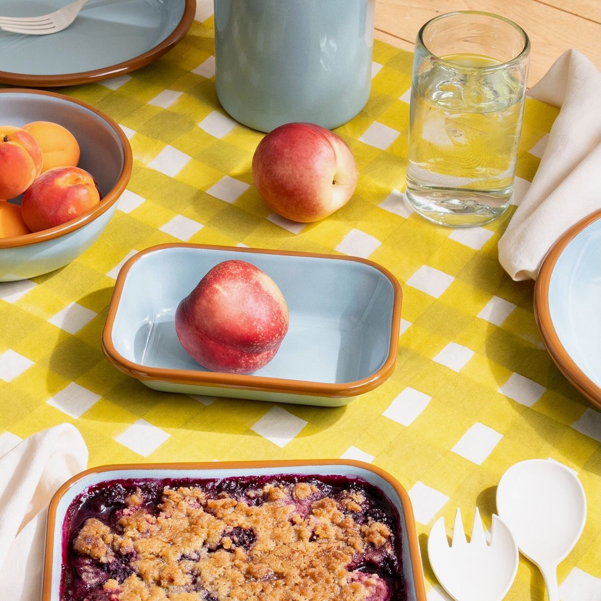 A small enamel baking dish with more enamel pieces, all in a robin's egg blue, with food and sitting on a table with a yellow crosshatch tablecloth.