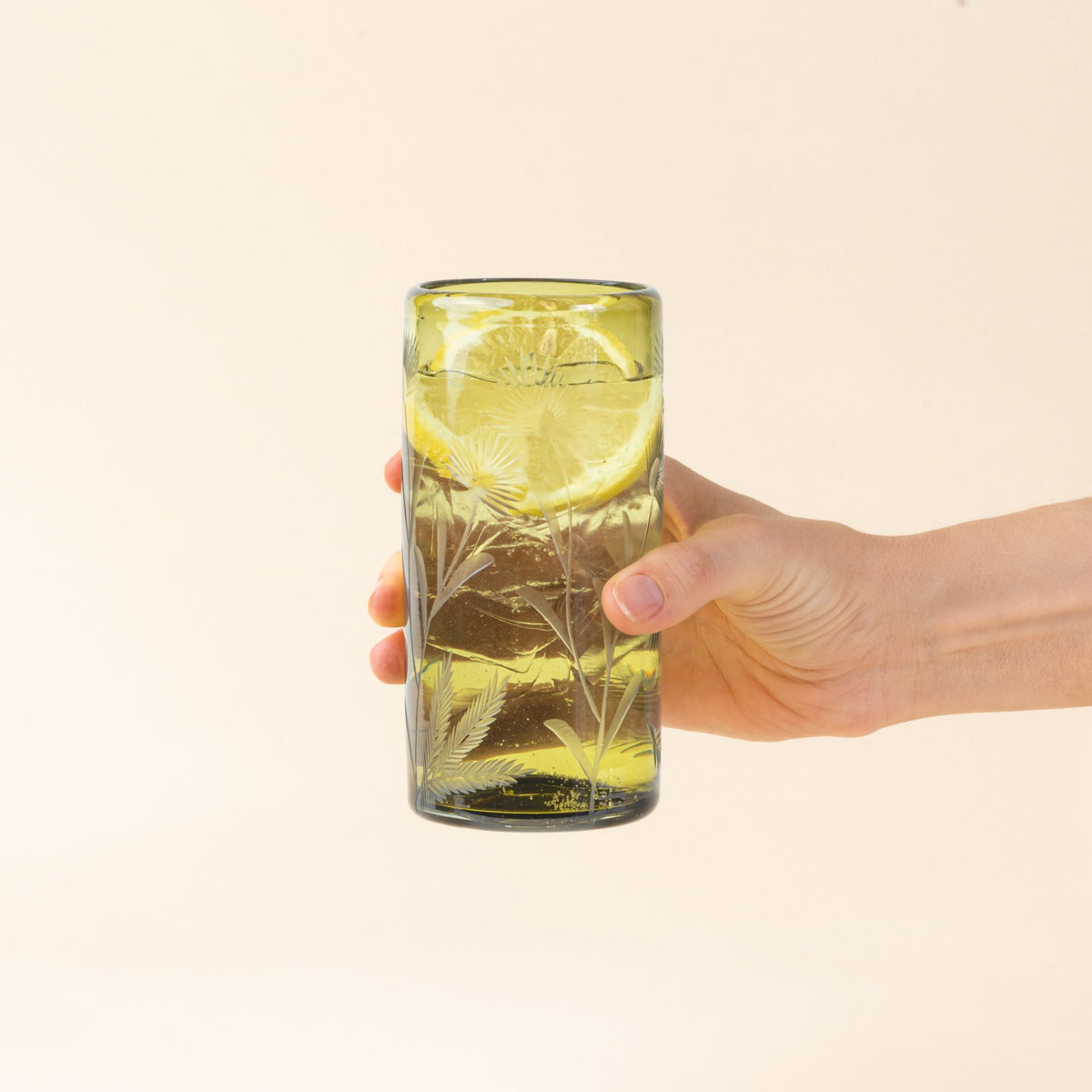 A hand holds out an olive cylindrical glass with delicate etched floral pattern, filled with water and a slice of lemon, standing upright on a neutral background.
