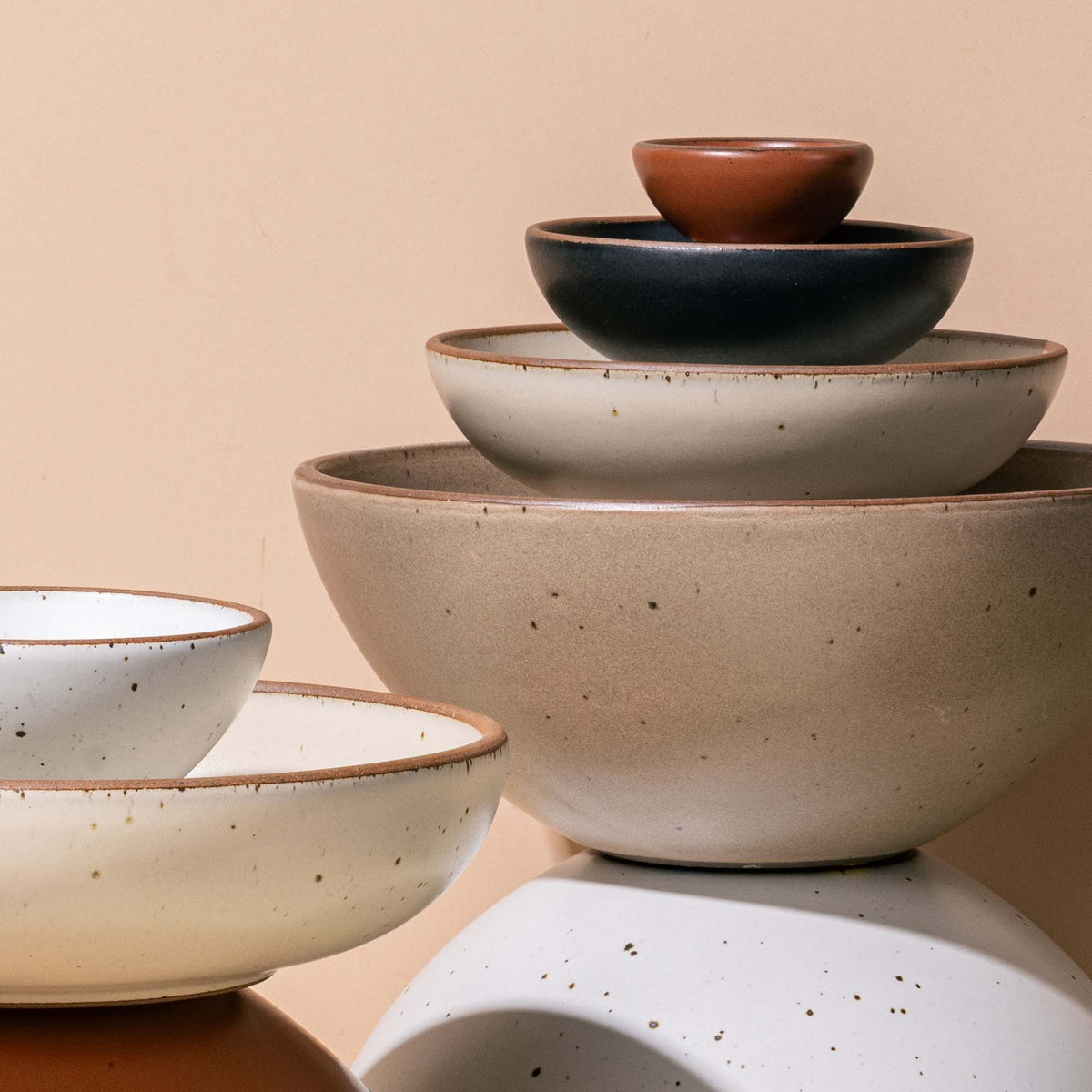 An artful stacking of ceramic bowls that range in sizes big to small and colors including, a warm tan brown, an off-white, graphite, and terracotta.