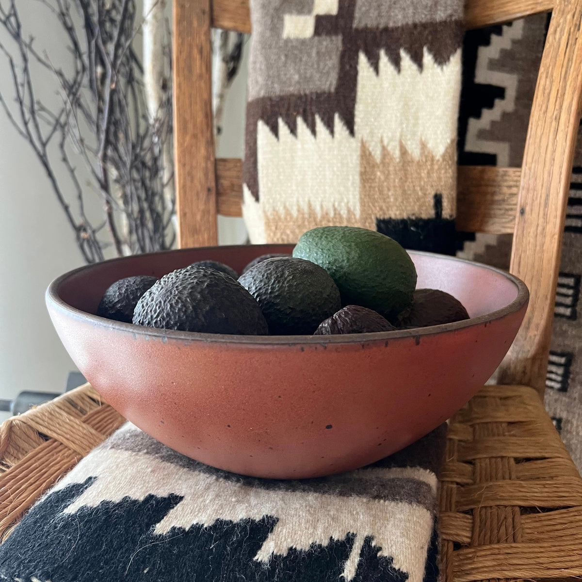A large terracotta ceramic bowl filled with avocados, set on a woven chair with a patterned textile backdrop.