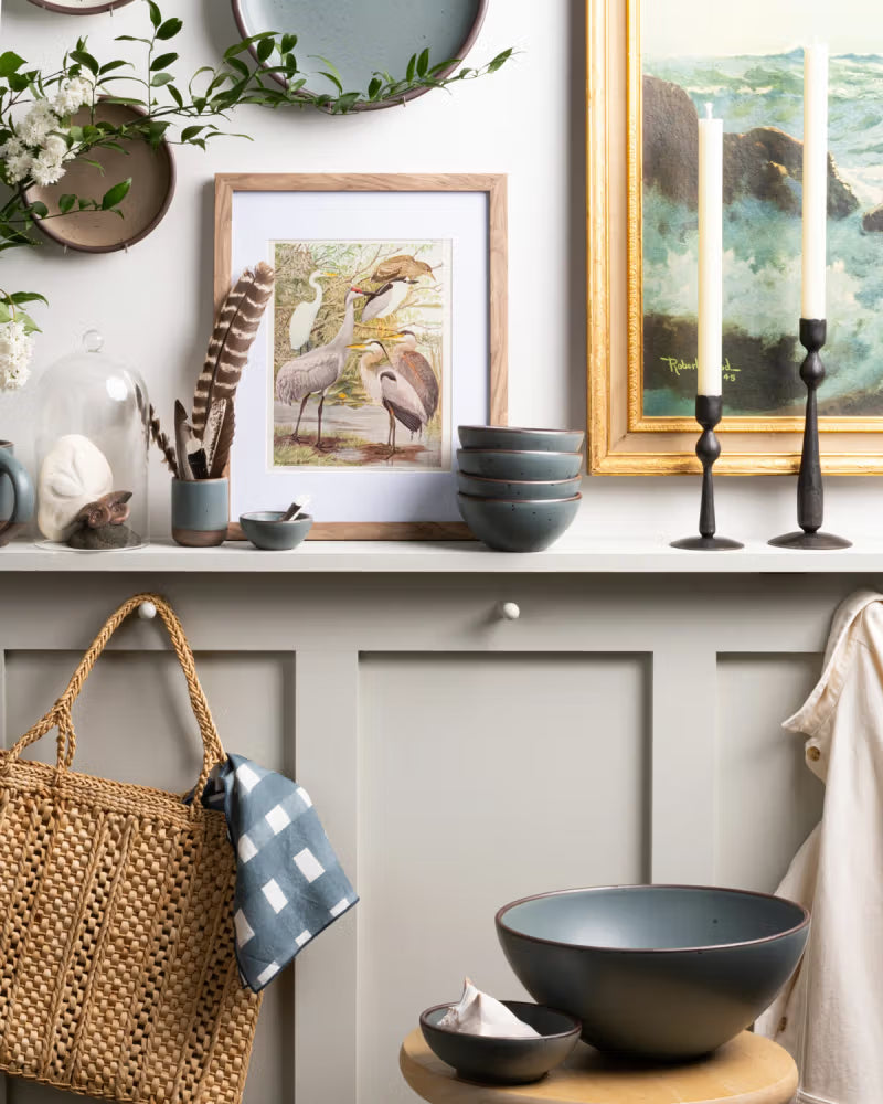 A calming entry shelf styled with ceramic bowls in a blue-grey color, a woven basket hanging on a hook, iron candlesticks, hanging art frames, and more.